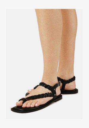 Picioare purtând sandale plate negre din piele împletită, cu barete cu cataramă, pe un fundal alb.