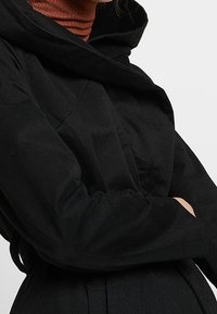 Musta päällystakki, jossa on huppu ja pitk ät hihat, vyöllä varustettuna. Kankaan pinta on sileä ja design on puhdas ja minimalistinen.