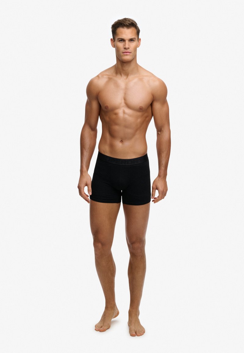 Homme musclé debout pieds nus, portant un boxer noir, faisant face à un fond blanc uni.