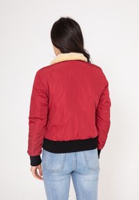 Veste bomber rouge avec un col en fourrure beige, des poignets et une taille élastiques noirs. Tissu lisse avec une silhouette cintrée.