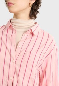 Pink striped button-up shirt with white and magenta vertical stripes, featuring a pocket and a soft texture, layered over a beige turtleneck.
