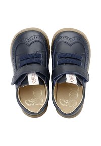 Zapatos infantiles de color azul marino con diseño en forma de corazón, parte superior de material sintético, cinta de velcro y suelas de goma beige. Los detalles de brogue adornan los zapatos.