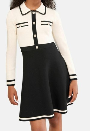 Mujer vistiendo un vestido de manga larga en blanco y negro con botones de perla, bolsillos delanteros y ribete a rayas en el cuello, los puños y el dobladillo.