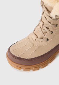 Nærbilde av beige og brune vinterstøvler med gummisåle, tekstillisser, sømdetaljer og fleecefôr synlig øverst.