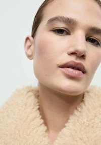 Primo piano di un volto con pelle liscia, trucco leggero e labbra piene. Indossa un colletto in shearling beige e testurizzato. Capelli castani naturali.