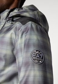 Chaqueta con capucha en tonos verdes y grises con un patrón de cuadros, que presenta un panel negro en el hombro y un parche con el logotipo bordado de EA7.