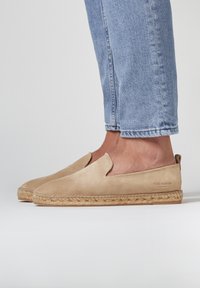 Espadrilles en suède beige à enfiler, avec une semelle en jute et des découpes. Portées avec un jean bleu clair, mettant en avant une texture lisse et un design décontracté.