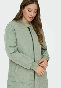 Viegli zaudēti, gaiši zaļš bomber jackets ar rāvējslēdzēju, ribētu aproču, sānu kabatām un gludu, spīdīgu tekstūru.