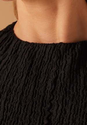 Gros plan sur le cou et le haut de la poitrine d'une personne portant un haut noir texturé à fronces avec un col rond.