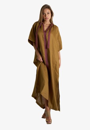 Donna che indossa un vestito lungo e largo color senape marrone con maniche ampie e rifiniture color granata, abbinato a tacchi aperti beige, in piedi su sfondo bianco.