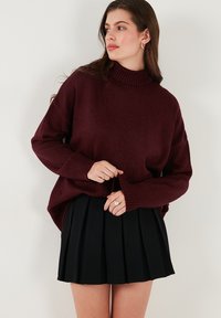 Burgundivörös kötött pulóver magas nyakkal, leengedett vállakkal és bordázott mandzsettákkal, fekete rakott mini szoknyával párosítva.