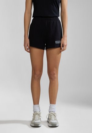 Vrouw gekleed in zwarte sportshorts, zwart mouwloos topje, witte enkelsokken en grijze hardloopschoenen, staand tegen een grijze achtergrond.