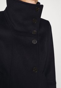 Abrigo de lana azul marino oscuro con un cuello alto, que presenta cinco botones negros y una ligera textura. Corte entallado con un diseño limpio y sin patrones visibles.