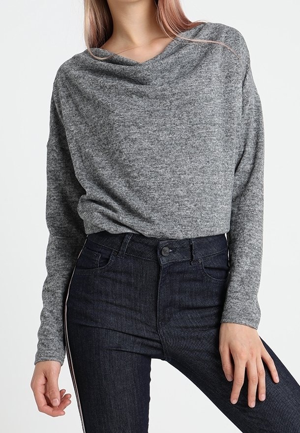 Femme portant un haut en maille gris à manches longues et un jean taille haute foncé avec une bande blanche sur le côté, posant avec une main sur la hanche.