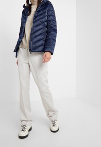 Marinblå puffjacka med huva, med ett diagonalt kviltat mönster, dragkedjefickor och en figurnära design. Matchas med krämfärgade vida byxor och vita stövlar.