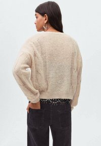Beżowy sweter dziany z detalami w warkocz, okrągłym dekoltem i luźnym krojem, zestawiony z ciemnymi dżinsami z dekoracyjnym paskiem.