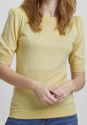 Vrouw met lichtbruin haar die een gele korte mouwen trui en blauwe jeans draagt, handen voor zich geklemd tegen een effen achtergrond.