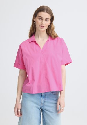 Donna con lunghi capelli castano chiaro, che indossa orecchini a cerchio d'argento, una camicia rosa acceso a maniche corte con colletto e jeans azzurri.