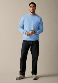 Lichtblauwe sweatshirts met geribbelde manchetten en zoom, gecombineerd met donkere jeans en grijze sneakers. Model staat met de handen gevouwen, op een neutrale achtergrond.