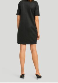 Schwarzes Kunstlederkleid mit kurzen Ärmeln und gerader Silhouette sowie Nahtdetail; getragen mit schwarzen High Heels, von hinten betrachtet.
