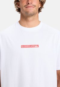 Hvid bomulds T-shirt med et rødt rektangulært logo med teksten "QUIKSILVER" og et lille grafik, korte ærmer og en rund halsudskæring.