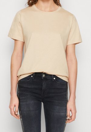 Persona que lleva una camiseta de manga corta beige metida en unos pantalones vaqueros ajustados oscuros con franjas laterales de la marca sobre un fondo blanco.