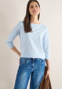 Femme portant un haut à manches trois-quarts rayé bleu clair et blanc et un jean bleu, tenant une veste marron texturée à la main.