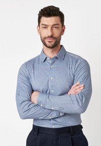 Man met baard en kort haar, gekleed in een blauw en wit gestreepte overhemden met knopen, met de armen gekruist, staande tegen een effen witte achtergrond.
