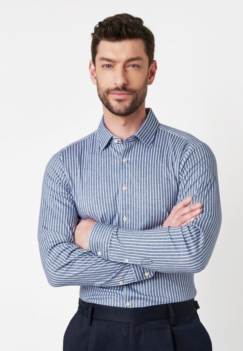 Man met baard en kort haar, gekleed in een blauw en wit gestreepte overhemden met knopen, met de armen gekruist, staande tegen een effen witte achtergrond.