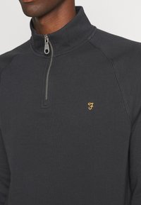 Primer plano de un sudadera de color gris oscuro con cremallera de un cuarto, con una pequeña "F" bordada en amarillo en el pecho, llevada por una persona.