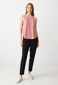 Donna che indossa un top senza maniche rosa, pantaloni neri su misura eSneakers bianche, in piedi contro uno sfondo bianco.