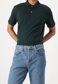 Muž oblečený v tmavě zeleném polo tričku s krátkým rukávem, které má zastrčené do světle modrých džín, s rukama za zády.