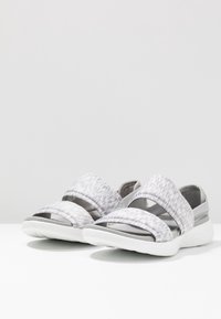 Scarpe sportive slip-on grigie e bianche con fasce incrociate, tessuto testurizzato e suola bianca imbottita. Design minimalista e costruzione leggera.
