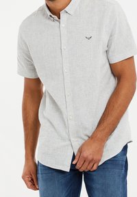 Camisa de botón de manga corta, de color gris claro, en una tela texturizada, con un logo bordado sutil en el pecho y un dobladillo redondeado.