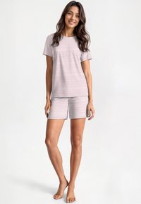 Young woman smiling, wearing white short-sleeve pajama set with thin red horizontal stripes, standing barefoot against plain background.