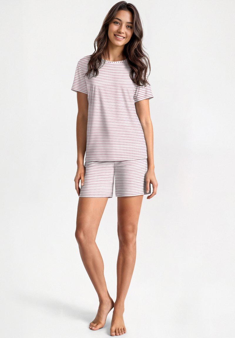 Young woman smiling, wearing white short-sleeve pajama set with thin red horizontal stripes, standing barefoot against plain background.