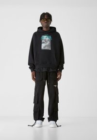 Melnā kapuca ar grafisku zīmējumu, kangaru kabatu un auklu kapuci; kopā ar melnajām kravas biksēm, kurām ir vairāki kabatas un logo.