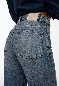 Sinisen denim-kankaan farkut, joissa on korkea vyötärö, suorat lahkeet ja nahkalamina, jossa on logo takavyötäröllä. Ei näkyvää kulutusta tai koristeita.