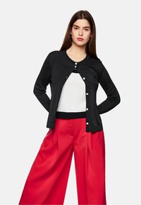 Schwarze Strickjacke mit rundem Ausschnitt, versehen mit silbernen Knöpfen. Getragen über einem strukturierten weißen Oberteil und kombiniert mit roten weit geschnittenen Hosen.