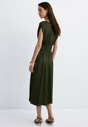 Femme portant une robe midi vert olive avec taille froncée et manches courtes, talons noirs à brides, et bracelets dorés, debout de profil.