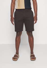 Shorts marron foncé en coton, avec une coupe classique et une longueur au-dessus du genou, accompagnés de sandales beiges à boucle.