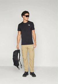 Melna īsām piedurknēm T-kreklu ar baltu logotipu, haki krāsas kargo bikses ar rāvējloku kabatām, melnu mugursomu un melnām kurpēm. Cirtaini mati, saulesbrilles.