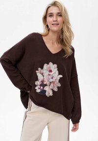 Brązowy, luźny sweter z dekoltem w kształcie litery V, ozdobiony kwiatowym nadrukiem. Sweter ma ściągacze na rękawach i zaokrąglony dół, w połączeniu z kremowymi spodniami.