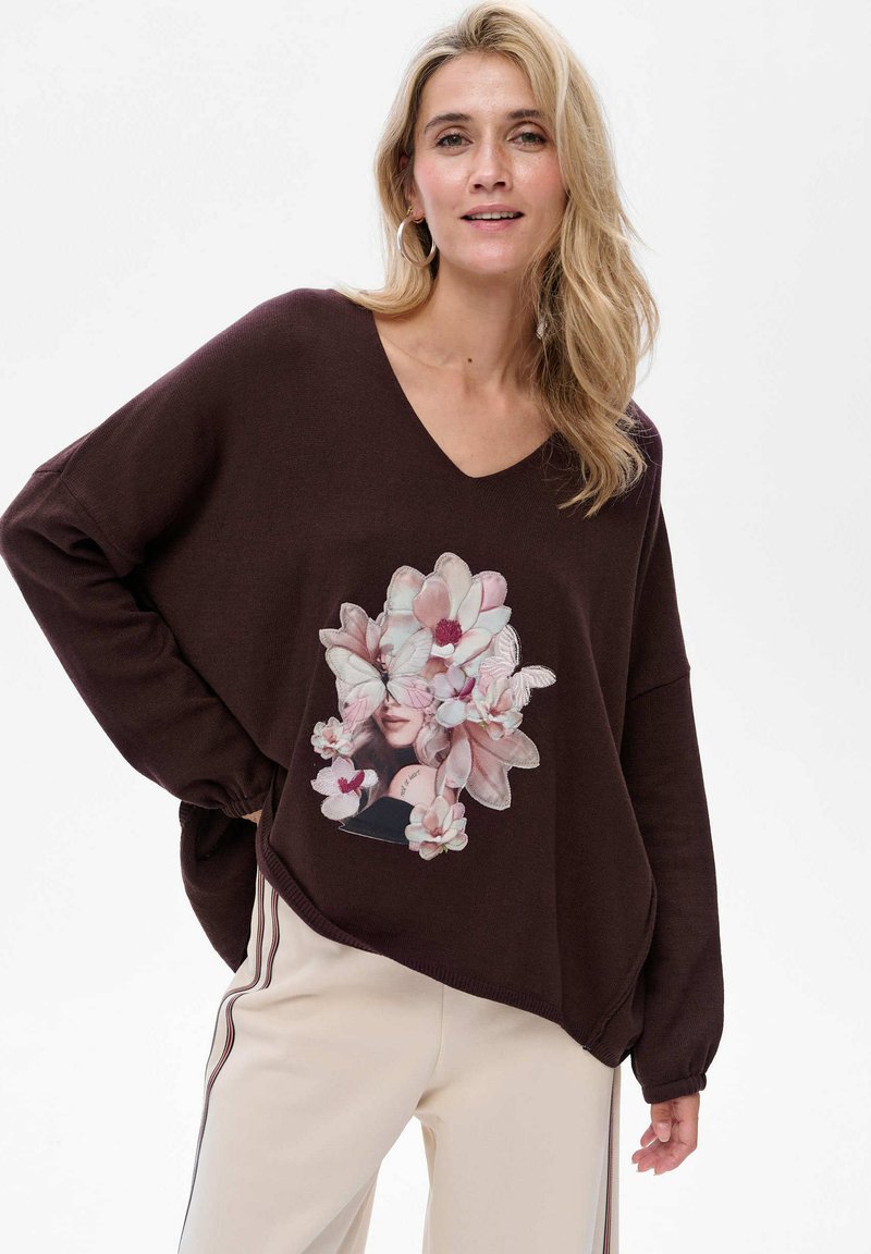 Brązowy, luźny sweter z dekoltem w kształcie litery V, ozdobiony kwiatowym nadrukiem. Sweter ma ściągacze na rękawach i zaokrąglony dół, w połączeniu z kremowymi spodniami.