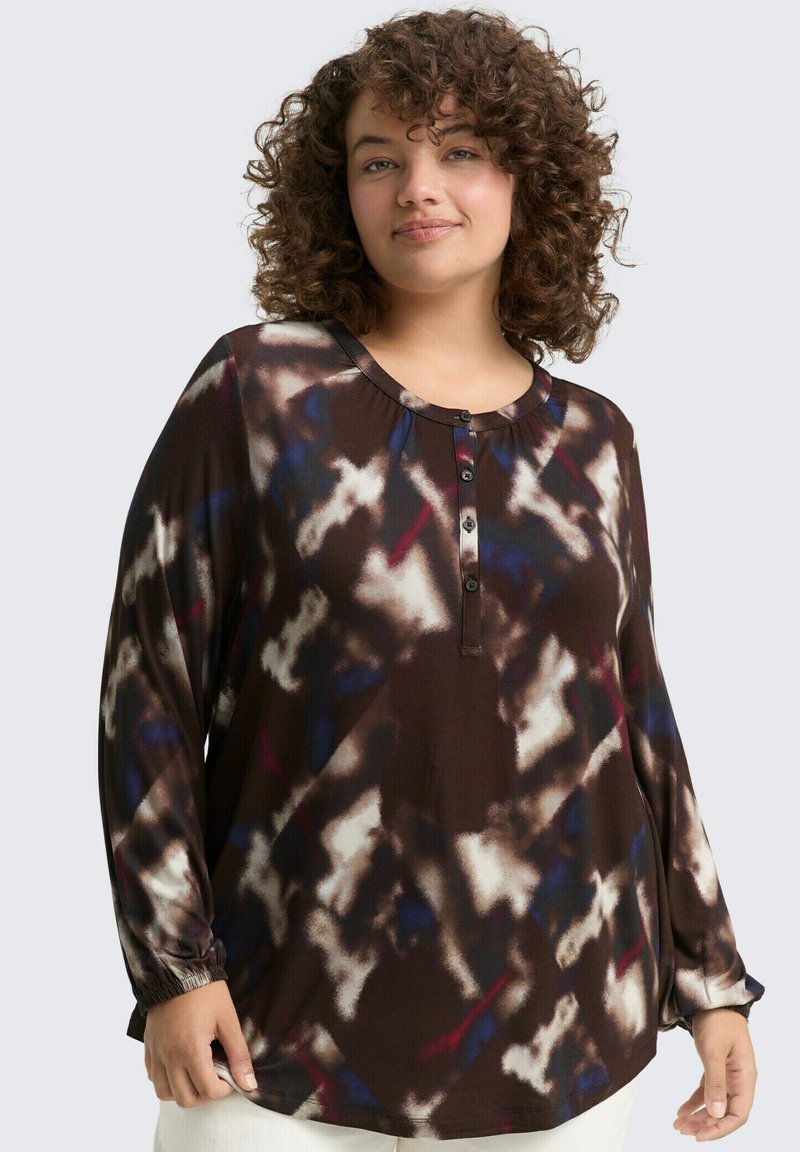 Glückige vrouw met krullend haar, gekleed in een blouse met lange mouwen, knopen en een abstract patroon in donkere tinten met witte en blauwe accenten.