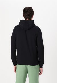 Hanorac negru cu o textură moale, având o glugă mare și manșete cu nervuri. Modelul îl asortează cu pantaloni de culoare verde deschis.
