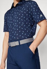 Námořnické polo tričko s geometrickým vzorem světle modrých trojúhelníků, doplněné o šedý pásek. Tkanina působí hladce s jemnou texturou.