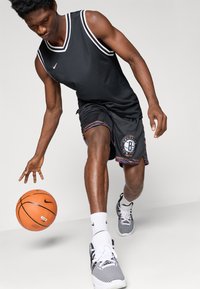 Top de desporto preto com riscas brancas, combinado com calções pretos com uma bainha colorida e o logótipo de Brooklyn. Bola de basquetebol laranja em movimento.