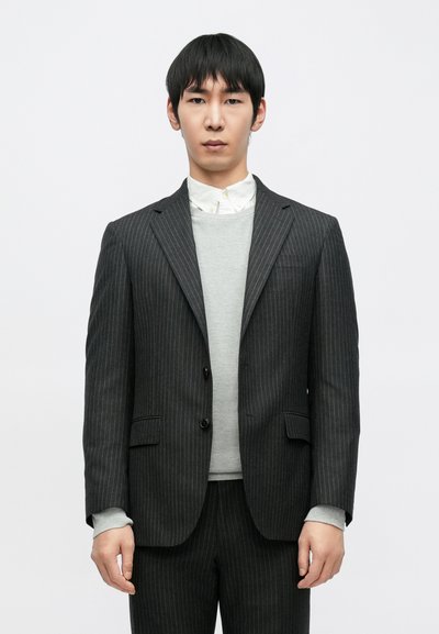 Tamnosivi odijelo s bijelim prugama s odgovarajućim hlačama, slojevito preko svijetlosivog džempera i bijele košulje s ovratnikom. Jednostavan, krojeni dizajn.