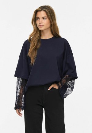 Kvinde iført en navyblå oversized top med gennemsigtige blonder lange ærmer, kombineret med sorte bukser, stående med den ene hånd i lommen.